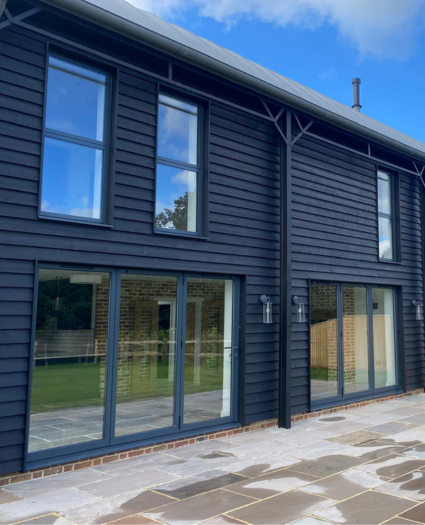 Modern two-storey house with dark timber cladding and large windows; ground floor features long sliding glass doors opening onto a stone patio and green lawn; part of Glaze Ltd project