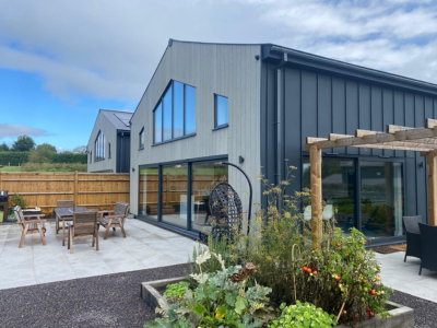Modern grey-clad house with angled roof and large floor-to-ceiling windows; outdoor patio with wooden dining table, benches and hanging egg chair; landscaped garden with small trees and shrubs in front; part of Glaze Ltd project