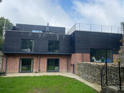 Two-storey modern house with brick lower level and dark cladded upper level with large windows; upper floor has balcony with glass balustrade; green lawn and gabion stone wall walkway in front; part of Glaze Ltd project