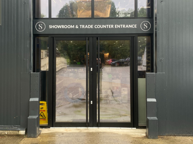 Showroom and trade counter entrance to dark corrugated metal building with double glass doors and signage; caution wet floor sign visible; part of Glaze Ltd project