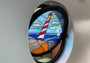 Circular stained glass window featuring lighthouse, sailboats and waves in red, blue and yellow colours set into a plain white wall; part of Glaze Ltd project