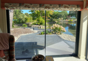 View from interior seating area through sliding glass door to landscaped patio with pond, plants and stone walkway; pillow and book on windowsill inside; part of Glaze Ltd project