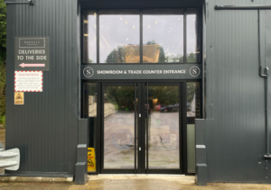 Trade counter and showroom entrance of dark corrugated metal building with double glass doors; signage indicates deliveries to the side and includes safety warnings; part of Glaze Ltd project