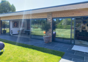 Modern single-storey building with timber slatted roof and large floor-to-ceiling glass doors opening onto dark slate patio and green lawn; potted olive tree outside and interior decorated with framed artwork and sofa; part of Glaze Ltd project