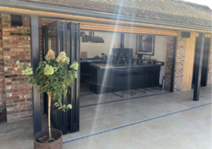 Outdoor covered bar area with brick column, open bi-fold doors and a long counter with high stools; potted tree on stone patio with sunlight streaming in; part of Glaze Ltd project