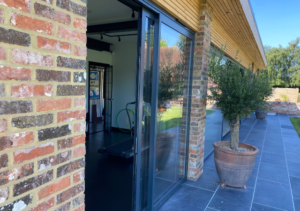 Open sliding glass door framed by a brick pillar leading to a home gym interior; grey tiled patio with potted tree and garden view; part of Glaze Ltd project