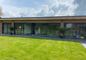 Single-storey building with timber roof and brick pillars, featuring floor-to-ceiling sliding glass doors; wide green lawn in front; part of Glaze Ltd project