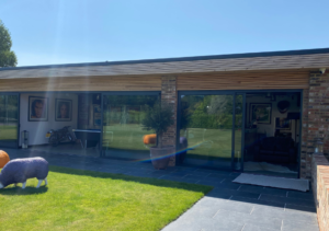 Modern garden outbuilding with brick pillar and timber roof overhang, large sliding glass doors; stone patio leading to green lawn with decorative sheep sculpture; interior lounge with framed pictures; part of Glaze Ltd project