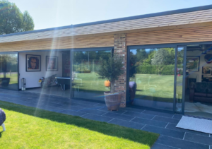 Modern single-storey structure with timber roof overhang, brick pillar and large sliding glass doors opening to an interior lounge area with framed pictures; grey tiled patio and green lawn; part of Glaze Ltd project