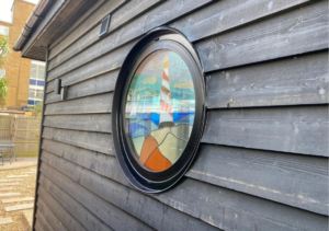 Circular stained glass window set into dark wooden cladding, featuring design of lighthouse and sailboats with blue and orange colours; part of Glaze Ltd project