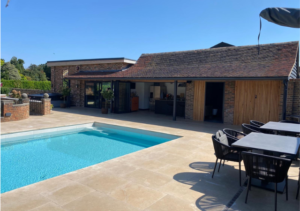 Outdoor swimming pool beside a single-storey wooden building with rustic tiled roof, sliding shutters and bar area; stone patio with dining table and chairs; part of Glaze Ltd project