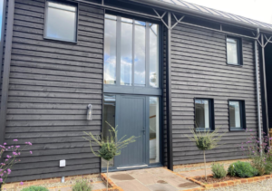 Front view of modern dark timber-clad building featuring a tall central glass entryway and grey front door, flanked by small topiary trees and landscaped flower beds; part of Glaze Ltd project
