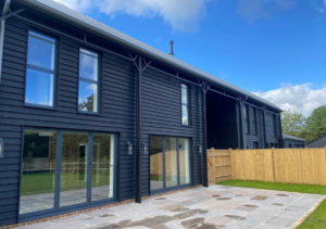 Modern dark timber-clad building with large sliding glass doors and tall windows above; stone patio and small lawn enclosed by wooden fence; part of Glaze Ltd project