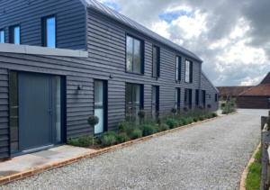Long dark timber-clad building with tall black-framed windows and grey door; gravel driveway lined with shrubs and topiary; barn structure visible in the distance; part of Glaze Ltd project