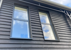 Close-up of two modern double-pane windows on dark wooden cladding, reflecting blue sky and clouds; part of Glaze Ltd project