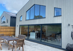 Modern grey timber-clad house with large sliding glass doors and expansive window above; stone patio with wooden table and chairs; part of Glaze Ltd project