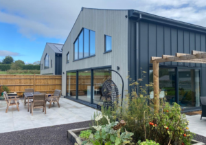 Modern grey-clad house with slanted roof and large black-framed windows and sliding doors; patio with outdoor dining set and hanging egg chair; wooden pergola and landscaped garden with flowering plants; part of Glaze Ltd project