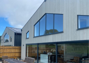 Modern grey-clad house with slanted roof and large black-framed windows and sliding doors; outdoor dining table and chairs on patio and wooden fence; part of Glaze Ltd project
