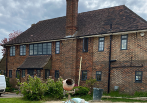 Two-storey traditional red brick house with pitched roof and chimney; multiple windows with dark frames; construction materials and cement mixer on lawn in front; part of Glaze Ltd project