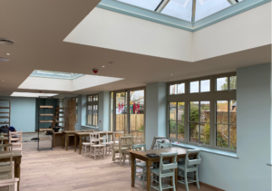 Bright dining area with rectangular skylights and large windows along one wall; wooden floors and white chairs with wooden tables; part of Glaze Ltd project