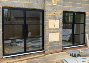 Building under construction with two large black-framed windows installed in a grey block wall; interior visible with insulation materials and wooden boards; construction debris on ground; part of Glaze Ltd project