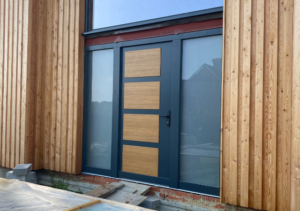 Modern front door with dark frame and three horizontal wooden panels set within vertical timber cladding, with full-height sidelight window and brick base and stone steps at entrance; part of Glaze Ltd project