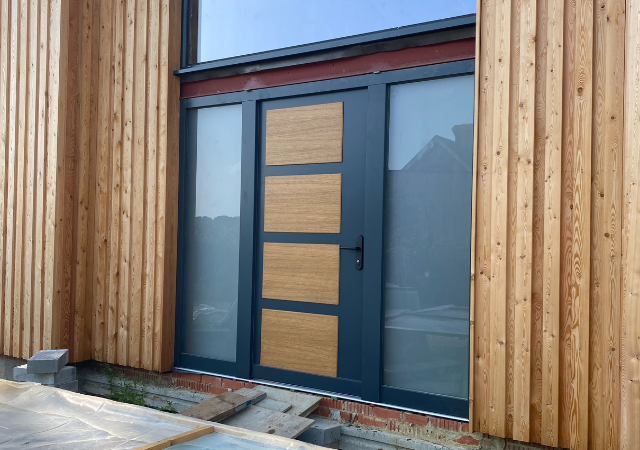 Modern front door with dark frame and three horizontal wooden panels set within vertical timber cladding, with full-height sidelight window and brick base and stone steps at entrance; part of Glaze Ltd project