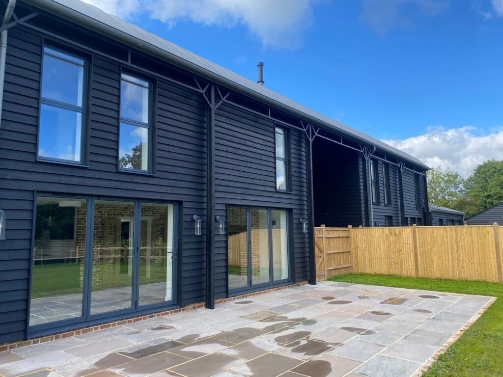 Modern dark wooden-clad two-storey house with multiple large sliding glass doors and tall windows; expansive stone patio and fenced garden; part of Glaze Ltd project.