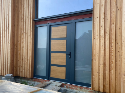 Modern front door with alternating wood and dark metal panels set in vertical timber cladding; construction materials at base; part of Glaze Ltd project.