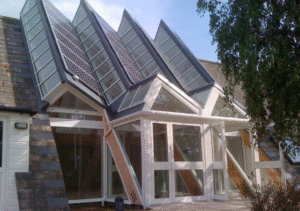 Modern glass conservatory extension featuring multiple slanted roof panels with solar panels, supported by white and wooden framing, attached to stone house; tree in foreground; part of Glaze Ltd project.