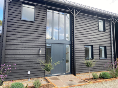 Modern dark wooden-clad building with tall central glass entryway, grey front door and rectangular windows; landscaped front garden with small trees and shrubs; part of Glaze Ltd project.